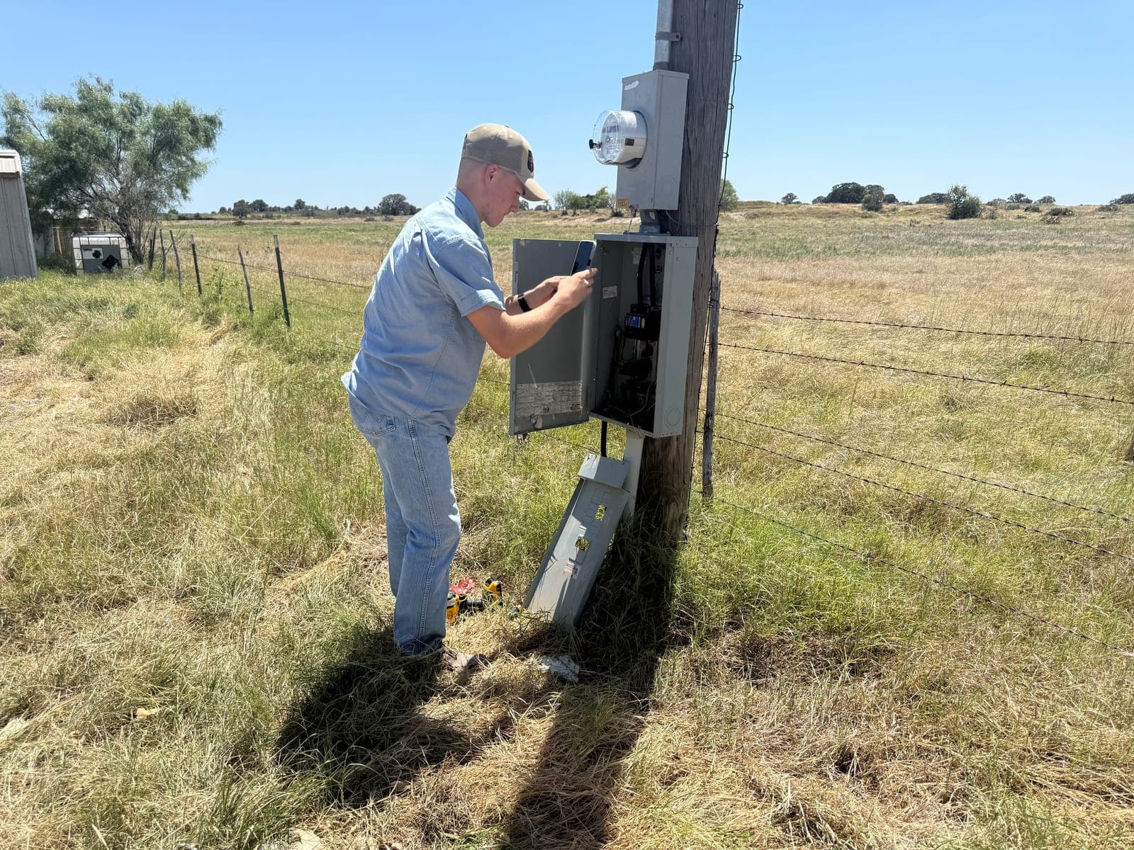 Technician opening an electrical panel mounted on a wooden pole below the meter, beside a barbed-wire fence