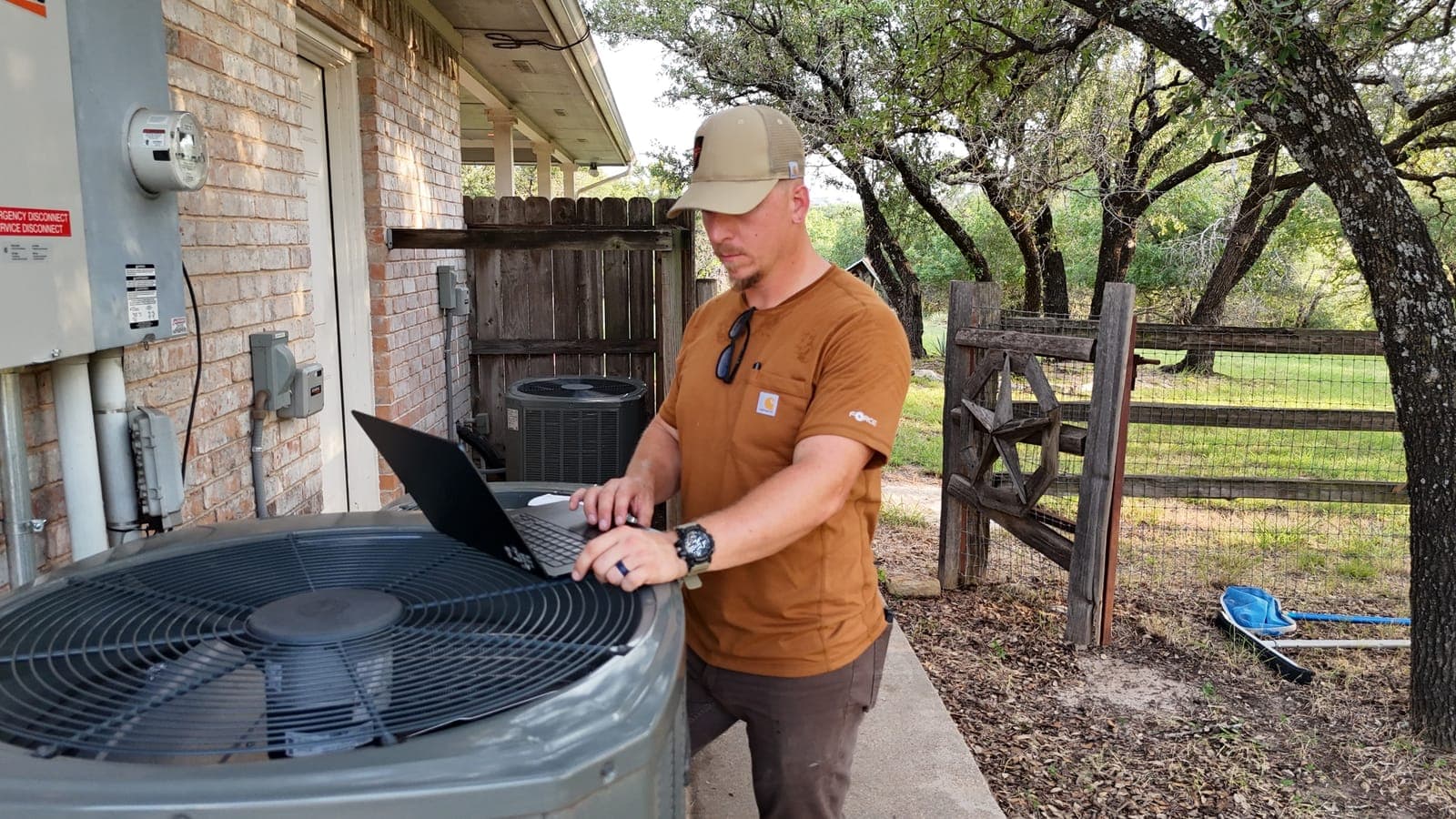 Technician working on a laptop on top of an outdoor AC condenser beside a brick house