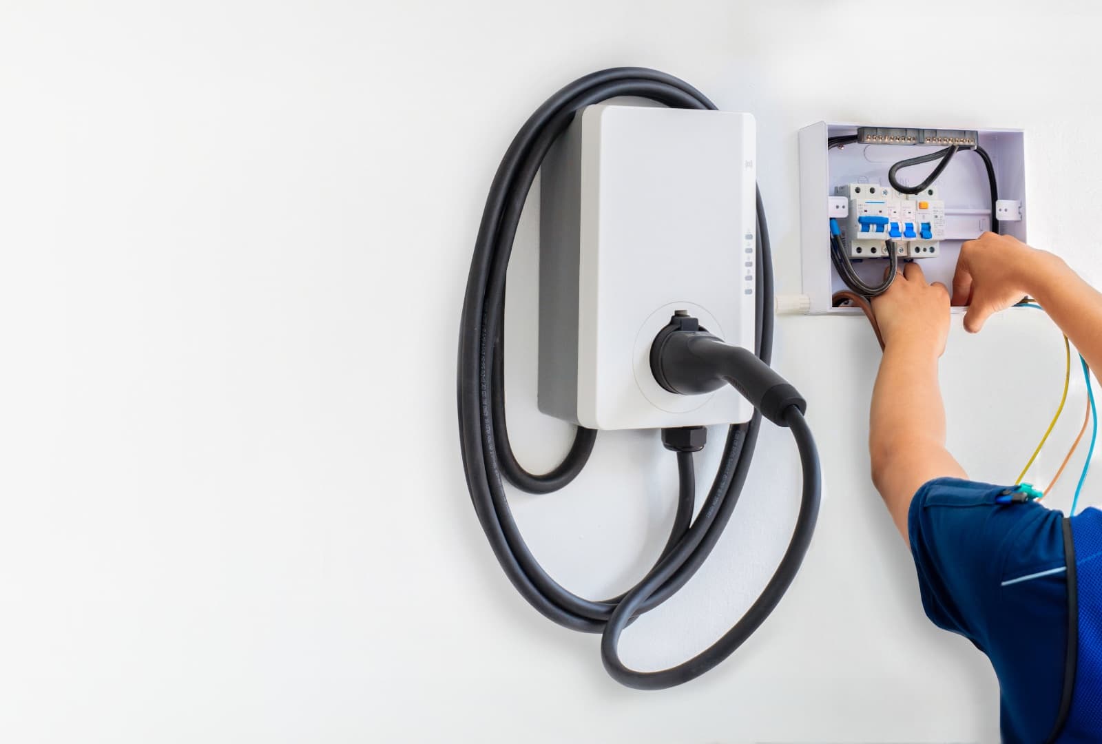 Wall-mounted EV charger with a coiled cable beside an open breaker box being wired by an electrician